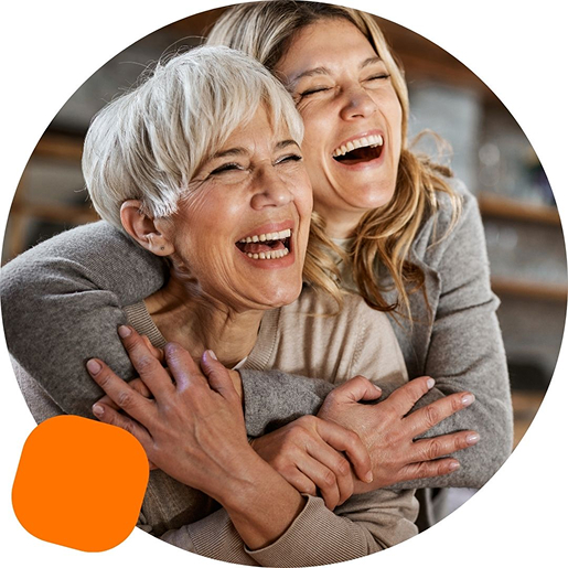 Two women laughing and hugging joyfully, one older with short gray hair and one younger with long blonde hair, both wearing cozy sweaters.