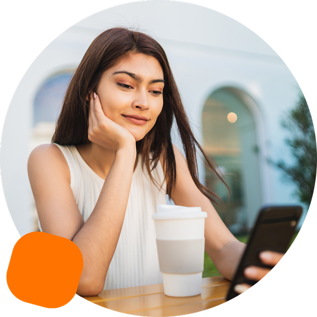 Young woman sitting at an outdoor table with a reusable coffee cup, looking thoughtfully at her phone.